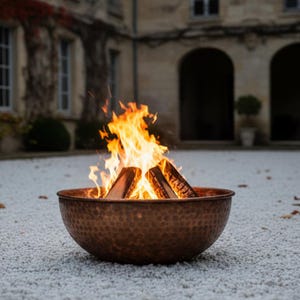 May include: A copper fire pit with a hammered texture, filled with burning firewood and bright orange flames. The fire pit is set on a bed of white pebbles, with a blurred background of a stone building.