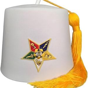 May include: A white fez hat with a yellow tassel. The hat features a colorful embroidered star emblem with various symbols. The tassel hangs down the side of the hat. The hat is a traditional style.