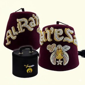 Op de afbeelding: Twee bordeauxrode fez-hoeden met gouden en kristallen versieringen, één met "Al Rai" en de andere met "Aires" in sierlijke letters. Een zwarte draagtas met een gouden embleem is ook aanwezig.