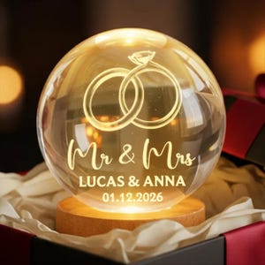 May include: A glowing glass sphere with a wedding ring design and the words "Mr & Mrs" above the names "LUCAS & ANNA" and the date "01.12.2026". The sphere sits on a wooden base, inside a gift box.