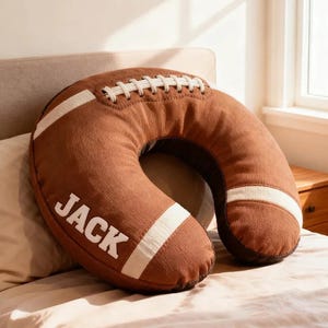 May include: Brown football-shaped neck pillow with white stripes and stitching. The word "JACK" is printed in white on the side of the pillow. The pillow is designed to provide neck support and comfort.