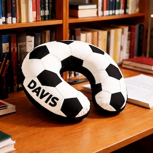 May include: A black and white soccer ball-patterned travel pillow with the name "DAVIS" printed in bold, black letters. The pillow is U-shaped and designed for neck support. It is resting on a wooden surface, with bookshelves in the background.