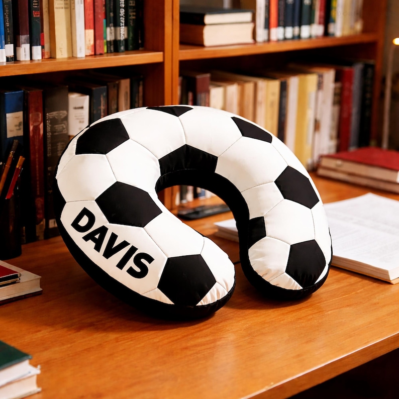 May include: A black and white soccer ball-patterned travel pillow with the name "DAVIS" printed in bold, black letters. The pillow is U-shaped and designed for neck support. It is resting on a wooden surface, with bookshelves in the background.
