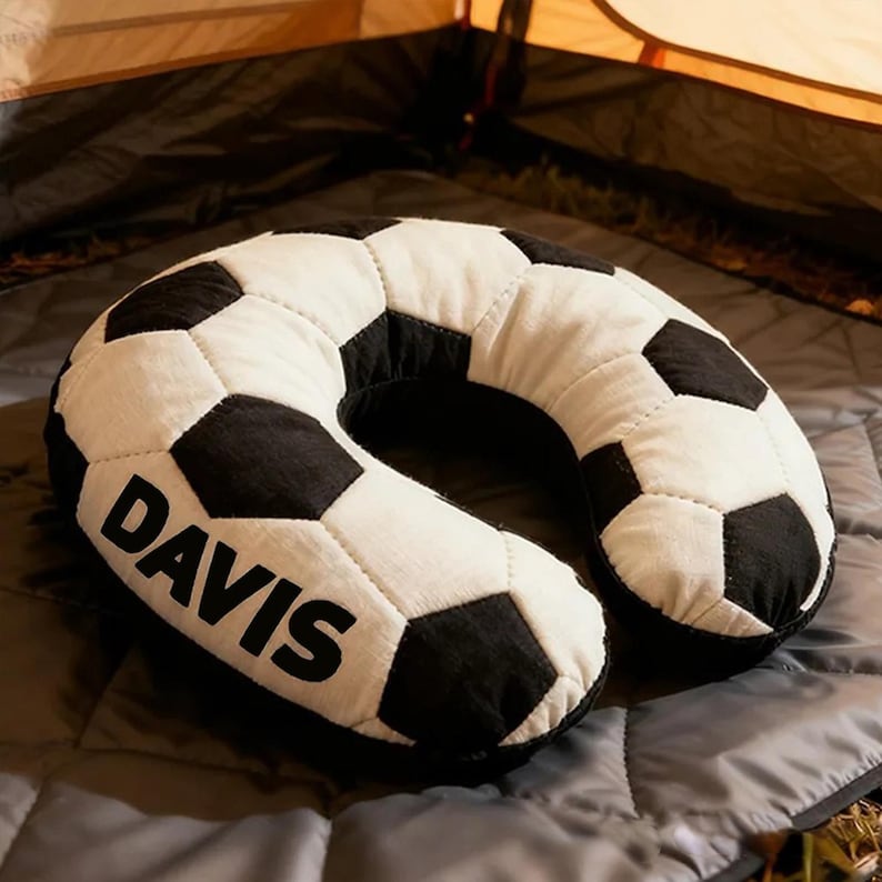 May include: A black and white soccer ball-shaped travel pillow with the name "DAVIS" printed in black. The pillow is on a gray blanket, with a tent in the background.