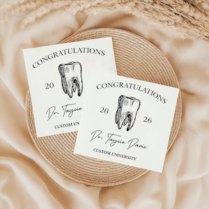 May include: Two white cards with the word "CONGRATULATIONS" printed at the top. Each card features a detailed illustration of a tooth and the name "Dr. Tayzie Davis" below. The cards are on a woven plate.
