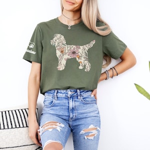 May include: Olive green t-shirt featuring a floral dog silhouette. The dog design is filled with pink, orange, and cream-colored flowers and leaves. The left sleeve has a paw print and the word "rainbow". The model is wearing blue jeans.
