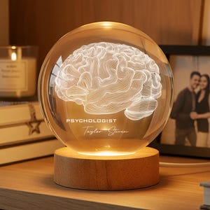 May include: A clear glass sphere with a 3D brain design, illuminated by a warm light. The sphere sits on a wooden base, with the words "PSYCHOLOGIST" and "Taylor Steven" etched on the glass. The lamp is on a wooden surface.