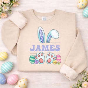May include: Beige sweatshirt with the name "JAMES" in blue letters. The design includes bunny ears, bunny feet, and Easter eggs. The bunny ears are light blue with purple polka dots. The bunny feet are light blue with pink paw pads.
