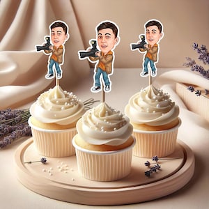 May include: Three cupcakes with white frosting and pearl sprinkles sit on a wooden tray. Each cupcake is topped with a cartoon cutout of a person holding a camera. The cupcakes are surrounded by lavender sprigs.