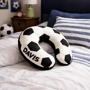 May include: A black and white soccer ball-shaped neck pillow with the name "DAVIS" printed on it. The pillow is on a bed with a blue and white striped pillow in the background. The pillow is designed to support the neck.