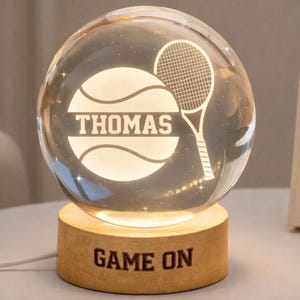 May include: A clear glass sphere with a tennis ball and racket design, illuminated from within. The name "THOMAS" is displayed on the ball. The sphere sits on a wooden base with the words "GAME ON".