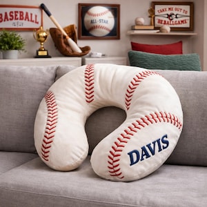 May include: A baseball-themed neck pillow in the shape of a horseshoe. The pillow is white with red stitching, mimicking a baseball. The word "DAVIS" is embroidered in blue. The pillow is resting on a gray couch.