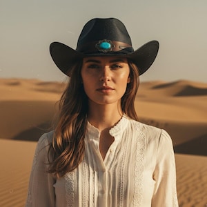 May include: A black cowboy hat with a turquoise stone detail. The hat features a brown leather band and a wide brim. The woman in the image is wearing a white blouse with lace detailing, set against a desert backdrop.