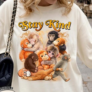 May include: Ivory-colored t-shirt featuring a graphic of several cartoon monkeys in various poses, with the text "Stay Kind" in an orange and yellow retro font. The shirt is from Comfort Colors.