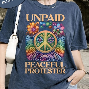 Puede incluir: Camiseta azul vaquero con las palabras "UNPAID PEACEFUL PROTESTER" en naranja. La camiseta presenta un símbolo de la paz con diseños florales y arcoíris coloridos. Las palabras "COMFORT COLORS" y "BLUE JEAN" están en un óvalo azul en la esquina superior izquierda.