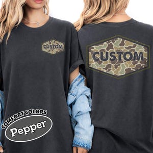 Puede incluir: Camiseta Comfort Colors gris oscuro con diseño de camuflaje y la palabra "CUSTOM" impresa en la parte delantera y trasera. La camiseta tiene mangas enrolladas y la palabra "Pepper" está impresa en la parte inferior.