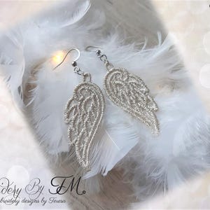 Puede incluir: Un par de pendientes plateados en forma de ala con intrincados detalles de encaje. Los pendientes se muestran sobre un lecho de suaves plumas blancas. La imagen incluye el texto "Embroidery By TM" y "Embroidery designs by Teresa".