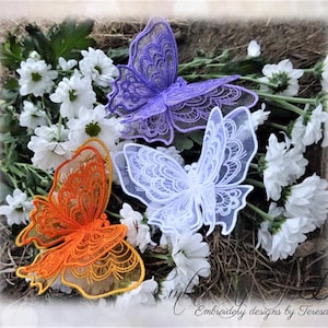 Puede incluir: Tres adornos decorativos de mariposas en naranja, morado y blanco, sobre flores blancas. Las mariposas tienen intrincados diseños de encaje. La imagen está etiquetada como "Embroidery designs by Teresa".