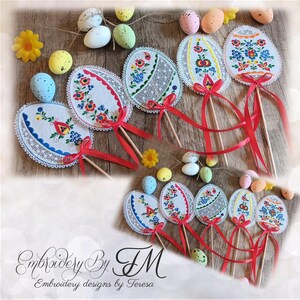 May include: Embroidered Easter egg decorations with floral designs, red ribbons, and wooden sticks. The eggs are white with colorful embroidery and lace trim. Decorative eggs and flowers are scattered around the eggs.