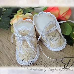 Può includere: Un paio di scarpine da neonato color avorio con delicati dettagli in pizzo floreale e nastri bianchi. Le scarpine sono ambientate su uno sfondo di rose e verde, con il testo "Embroidery By TM".