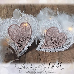 Könnte beinhalten: Zwei herzförmige Spitzenstickereien. Das äußere Herz ist weiß mit einem gewellten Rand, und das innere Herz ist staubrosa mit einem Blättermuster. Weiße Federn und der Text "Embroidery By FM" sind ebenfalls sichtbar.