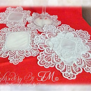 Könnte beinhalten: Weiße Spitzentischsets mit quadratischer Mitte und aufwendigen floralen Designs werden auf einer roten Oberfläche präsentiert. Die Tischsets sind um ein Weinglas herum angeordnet und zeigen ihre dekorative Wirkung. Das Bild enthält den Text "Embroidery By TM".