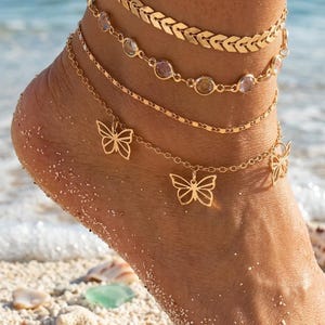 May include: A layered gold anklet set featuring a chevron chain, a chain with clear and purple stones, and a chain with butterfly charms. The anklets are worn on a foot on a sandy beach with the ocean in the background.