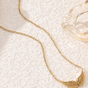 May include: A gold-colored necklace with a chain and a pendant shaped like angel wings. The wings are detailed with feather-like textures. The necklace is displayed on a textured white surface.