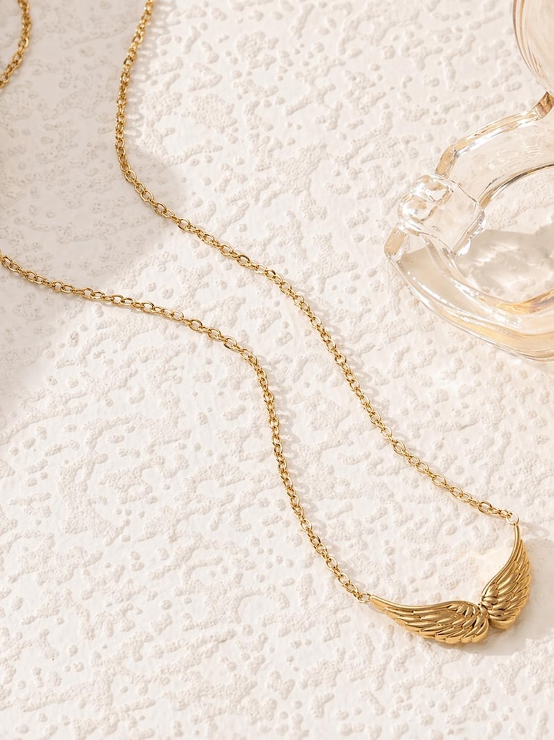 May include: A gold-colored necklace with a chain and a pendant shaped like angel wings. The wings are detailed with feather-like textures. The necklace is displayed on a textured white surface.