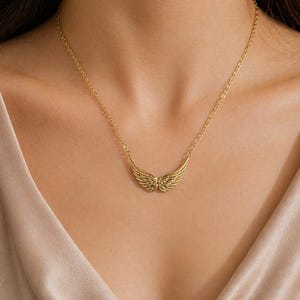 May include: A gold-colored necklace featuring a pendant shaped like angel wings. The wings are detailed with feather-like lines and are suspended from a delicate chain. The necklace is worn against a neutral-toned background.