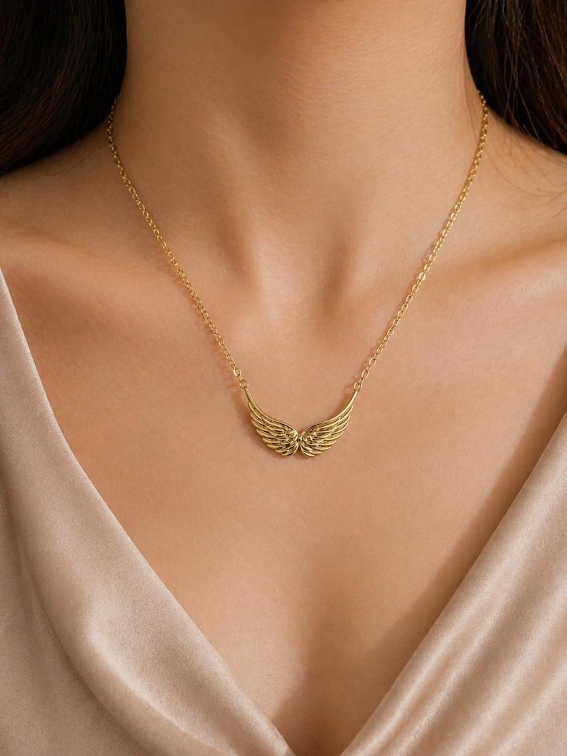 May include: A gold-colored necklace featuring a pendant shaped like angel wings. The wings are detailed with feather-like lines and are suspended from a delicate chain. The necklace is worn against a neutral-toned background.