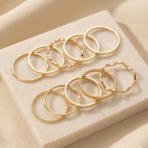May include: A collection of twelve gold-toned rings arranged in two rows. The rings feature various designs, including smooth bands, twisted rope textures, wavy edges, and notched patterns. Each ring is a simple circular band made of metal.