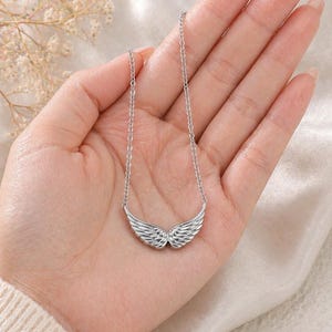 May include: A silver-colored necklace featuring a pendant shaped like angel wings. The wings are detailed with feather-like textures and are suspended from a delicate chain. The necklace is displayed on a person's hand.