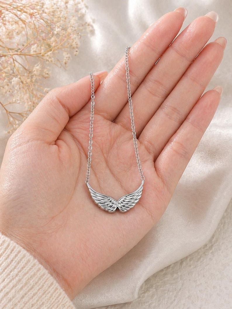 May include: A silver-colored necklace featuring a pendant shaped like angel wings. The wings are detailed with feather-like textures and are suspended from a delicate chain. The necklace is displayed on a person's hand.