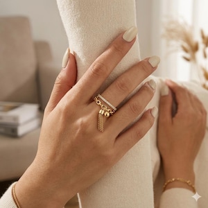 May include: A gold-toned ring with a rectangular top adorned with small, clear stones. A chain and tassel hang from the ring, adding a decorative element. The ring is on a person's finger, with a matching bracelet visible in the background.
