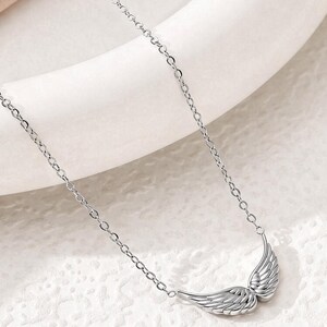 May include: A silver-colored necklace featuring a pair of detailed wing pendants. The wings are positioned side-by-side, with intricate feather detailing. The necklace chain is delicate and links to the wings.