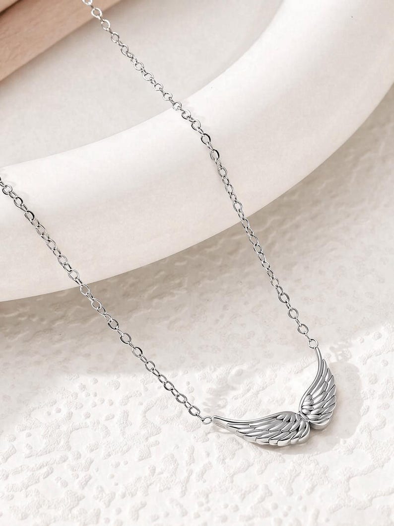 May include: A silver-colored necklace featuring a pair of detailed wing pendants. The wings are positioned side-by-side, with intricate feather detailing. The necklace chain is delicate and links to the wings.
