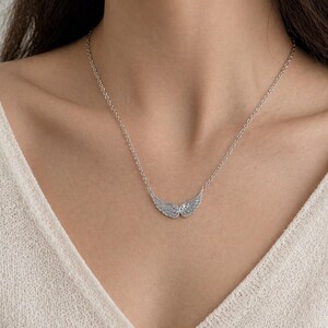 May include: A silver-colored necklace featuring a pair of detailed wing pendants. The wings are positioned horizontally, with delicate feather detailing. The necklace is on a delicate chain, and the background is a cream-colored top.