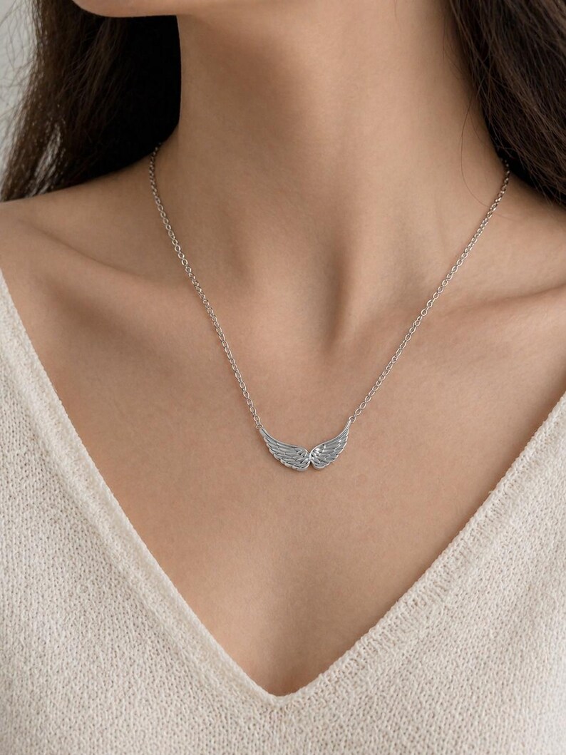 May include: A silver-colored necklace featuring a pair of detailed wing pendants. The wings are positioned horizontally, with delicate feather detailing. The necklace is on a delicate chain, and the background is a cream-colored top.