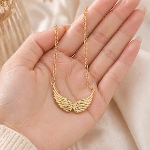May include: A gold-colored necklace featuring a pendant shaped like angel wings. The wings are detailed with feather-like lines. The necklace is displayed on a hand against a soft, neutral background.