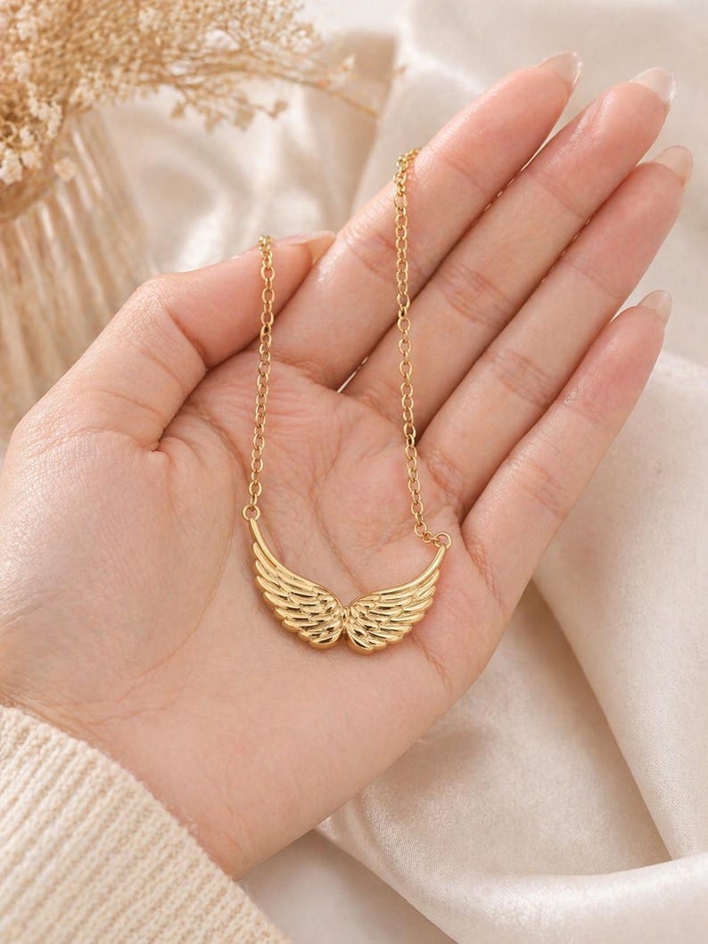 May include: A gold-colored necklace featuring a pendant shaped like angel wings. The wings are detailed with feather-like lines. The necklace is displayed on a hand against a soft, neutral background.