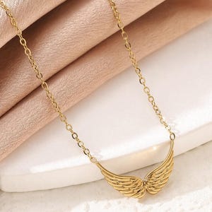 May include: A gold-colored necklace featuring a pair of stylized wings. The wings are detailed with feather-like lines and are connected to a delicate chain. The necklace is displayed on a light pink fabric.