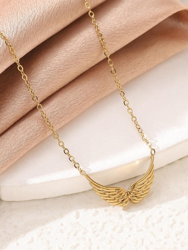 May include: A gold-colored necklace featuring a pair of stylized wings. The wings are detailed with feather-like lines and are connected to a delicate chain. The necklace is displayed on a light pink fabric.