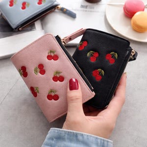 May include: A close-up of three small, zippered wallets in different colors. The wallets are light pink, black, and light blue. Each wallet has an embroidered cherry pattern. A hand is holding the pink and black wallets.
