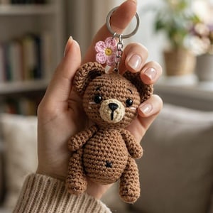 Könnte beinhalten: Ein brauner gehäkelter Teddybär-Schlüsselanhänger mit rosa Blumenakzent. Der Bär hat schwarze Augen und eine hellbraune Schnauze. Der Schlüsselanhänger wird von einer Person gehalten, die einen beigen Pullover trägt.
