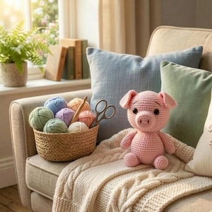 Könnte beinhalten: Ein rosa gehäkeltes Schwein sitzt auf einer cremefarbenen Decke. Ein Korb mit Garn in verschiedenen Pastellfarben, eine Schere und eine Häkelnadel befinden sich neben dem Schwein. Die Szene spielt sich auf einem beigen Sofa mit blauen und grünen Kissen ab.