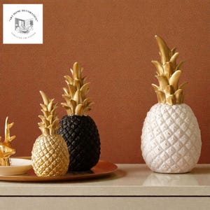 Puede incluir: Esculturas decorativas de piña en dorado, negro y blanco. Las piñas tienen superficies texturizadas y copas doradas. La imagen también incluye una figura de ciervo dorada y un plato. El fondo es de color marrón liso.