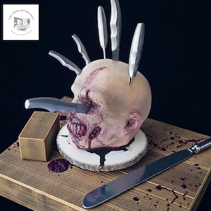 May include: A macabre Halloween decoration depicting a severed head with knives embedded in it. The head sits on a wooden slice, with a wooden block and a silver knife. Red liquid splatters enhance the horror theme.