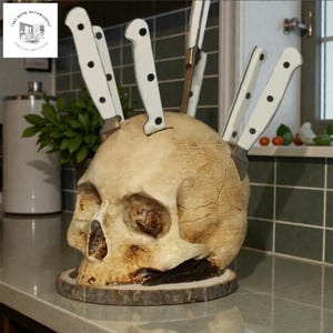 May include: A decorative skull-shaped knife holder with several knives inserted. The skull is beige with dark brown accents, and the knives have white handles with black dots. The holder sits on a wooden base.