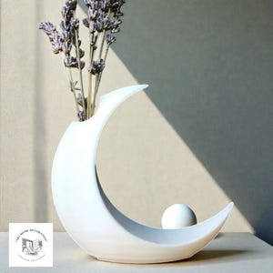 May include: A white ceramic crescent moon vase with a spherical accent. The vase holds dried lavender stems. The design is minimalist and modern, suitable for home decor. The vase is on a light-coloured surface.